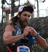 Un homme blanc barbu aux cheveux long portant une casquette de cyclisme et un gilet de course.