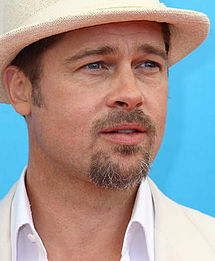 A Caucasian with light brown hair, blue eyes, and a mustache and short brown beard, in front of a turquoise background. He is wearing a white shirt and white hat.