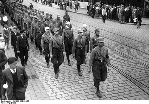 A black and white photograph of a column of Nazi brownshirts marching through the streets of Berlin.