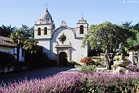 Ön planda mor çiçeklerle yuvarlak cephe ve her iki tarafta küçük çan kuleleri ile Adobe kilise