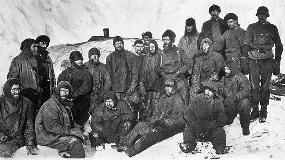 A group of men sitting closely packed together in heavy winter clothes and hats. Snow and ice surrounds them.