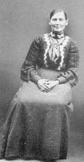 Photographie d'une femme de face, en robe, les cheveux tirés, assise et penchée vers la droite, les mains croisées sur les genoux.