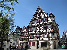 Um einen Platz angeordnet: eine Stele mit der Aufschrift „Place de Tarare“, Fachwerkhäuser und ein Brunnen