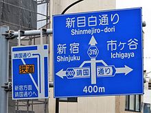 Japanese road sign directing away from a narrow roadway. Left board says "To Shinjuku, go ahead to Yasukuni-dori Ave.", and the ideograms written in the yellow rectangle mean "narrow street" Kappazaka intersection road sign.JPG