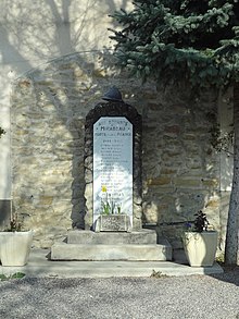 Das Kriegerdenkmal: eine einfache Stele, überragt von einem Adrian-Helm, vor dem Rathaus aufgestellt.  Im Putz des Rathauses wurde ein Mangel in Form eines Bogens gelassen, der ihn hervorhebt