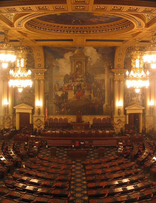 A view of a large, ornately-decorated room with several rows of curved desks, arranged in a semicircle. A large mural is visible on the wall at the far end of the room.