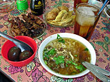 A selection of Indonesian food, including Soto Ayam (chicken soup), sate kerang (shellfish kebabs), telor pindang (preserved eggs), perkedel (fritter), and es teh manis (sweet iced tea)