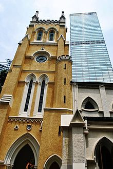 Sebuah menara gereja kuning bergaya Gothik yang terlihat dari bawah, dengan gedung pencakar langit bereksterior kaca di belakangnya.