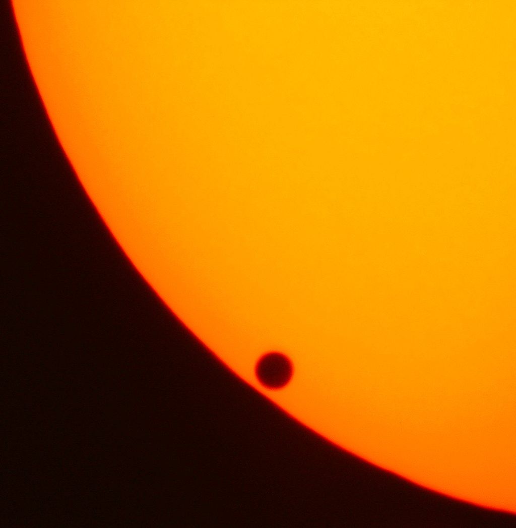 Venus appears as a black bubble on the edge of the Sun's disk, dimmed through filters to a dull orange.
