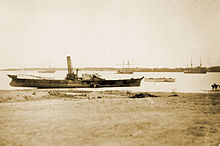 Photograph showing the hulk of a side-wheel steamship sitting very high in shallow water and heavily listing adjacent to a beach with other ships in the background