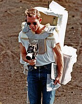Man around age 40 with sunglasses and a large backpack takes a photograph with a camera mounted on his chest