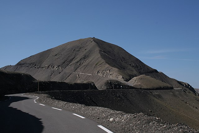 View from an asphalt road up to a pointed dark grey mountain peak with a surrounding road