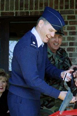 A profile photo of white man in a blue US Air Force uniform. He is facing the right, smiling, and wielding large scissors for a ribbon-cutting ceremony.