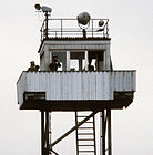 Auf einer Stahlrohrtragstruktur mit einer Leiter befindet sich eine kleine Kabine mit Fenstern nach allen Seiten, die von einem Balkon umgeben ist, auf dem zwei Männer in Uniform stehen und den Betrachter durch ein Fernglas betrachten. Ein dritter Mann ist in der Kabine. Auf dem Flachdach des Turms befinden sich Scheinwerfer und ein Geländer um die Außenkante des Dachs.