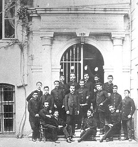 Photographie en noir et blanc d'une vingtaine d'étudiants en uniforme scolaire se tenant debouts et assis devant l'entrée d'une école.