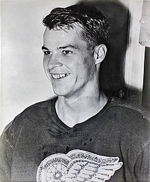 Photo noir et blanc d’un homme, jeune, souriant, de 3/4 face, portant un pull foncé avec une roue ailé sur le devant