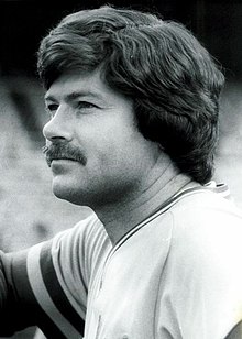 A man wearing a light baseball jersey with wide stripes around the sleeve openings