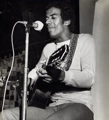 Photographie en noir et blanc d'un homme assis qui joue de la guitare.