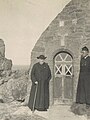 Duas pessoas em batinas pretas, a da esquerda carregando uma bengala e um chapéu, estão perto de uma capela de pedra (vemos uma porta de madeira)