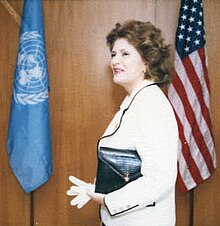 A woman holding a purse and gloves, standing between the UN and US flags