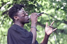 Sumney wearing a baggy black t-shirt and sunglasses. He is holding a wired microphone close to his mouth with his right hand, and making a loose pointing gesture with his left. He is outdoors in the summer, and the background primarily consists of thick tree cover.