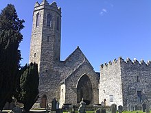 Photo of east elevation showing three contiguous elements, narrow tall octagonal tower left, gable with stained glass window centre, low square-plan tower right.