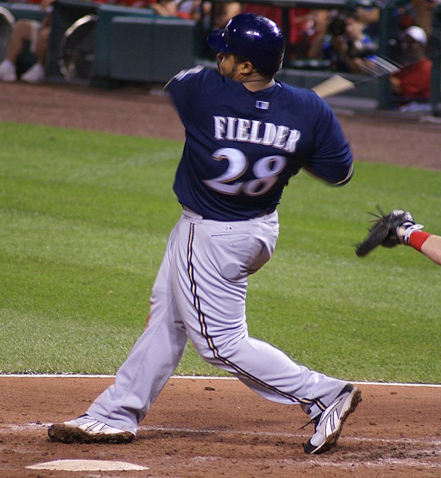 A man in a navy baseball jersey and batting helmet with gray pants