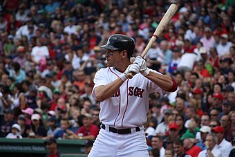 A man in a white baseball uniform with "RED SOX" on the chest and a batting helmet stands in a left-handed batting stance with a large crowd in the background