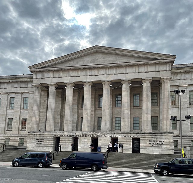 Exterior of a large building made of grey stone. Columns mark the entrance and a wide set of stairs lead up to the columns. Three cars are parked in front of the building. The sun peeks through the cloudy sky above. Several people are walking toward the entrance.