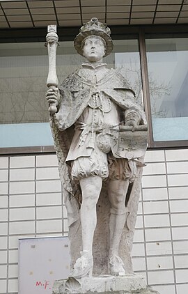 A stone statue of Edward VI. Edward wears a crown and holds a parchment in his left hand and a sceptre in his right hand. The king stands on a stone plinth.