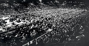 thousands of spectators in a large arena look towards a man standing behind a podium on a platform