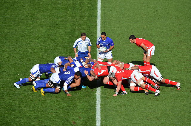 A player holds a ball in front of two opposing groups of eight players. Each group is crouched and working together to push against the other team.
