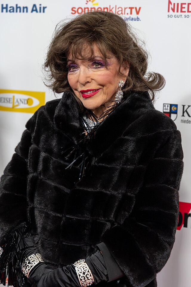 Joan Collins in einem dunklen Pelzmantel vor Werbewand