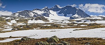 Fladt landskab med snemarker i forgrunden og bjerg i baggrunden med en gletscher omgivet af lodrette klippeflader.