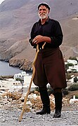 Cretan shepherd dressed in traditional Cretan outfit posing for a photo. He wears: kefalomantilo, black long-sleeve shirt, kilotes/vraka breeches tucked into stivania, and is also leaning on a katsouna.