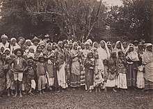Indische Indentur-Arbeiter. Schwarz-Weiß-Bild von Männern, Frauen und Kindern in Saris und teilweise mit Kopftüchern vor tropischer Vegetation.