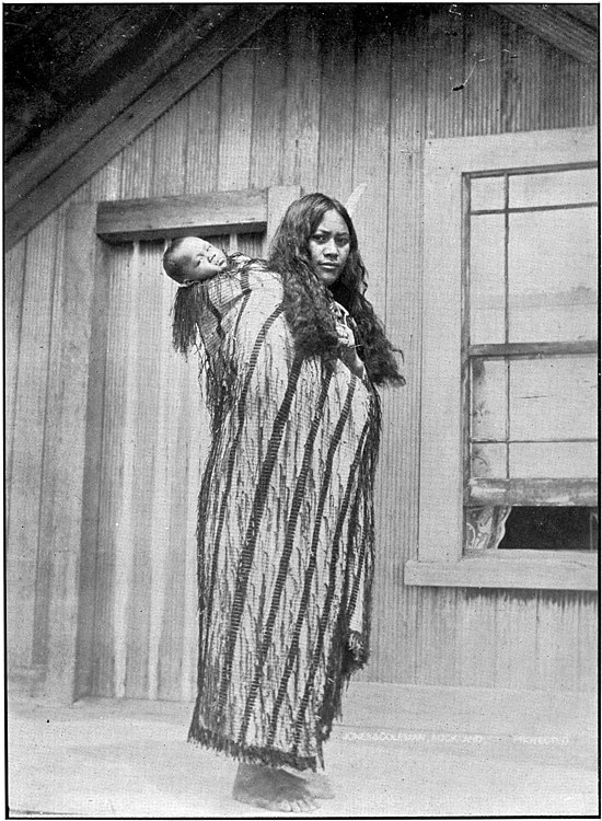 "A Māori women with a baby on her back wearing a cloak, outside a house with a door and partially opened window"