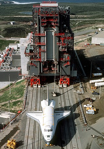 1985 photo of Space Shuttle Enterprise (OV-101) moving toward the shuttle assembly building at Vandenberg Space Launch Complex-6 aboard its specially designed Cometto 76-wheel transporter. In the background are the payload changeout room and the payload preparation room.
