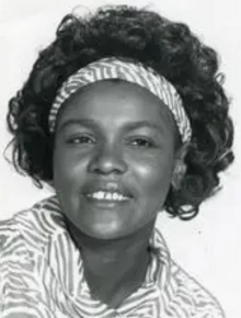 A smiling Black woman with hair in a curly bouffant style; she is wearing a print top and a matching headband