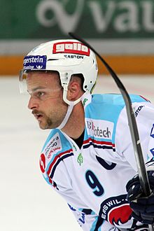 Kleurenfoto van een man, zijn hoofd in profiel, naar links kijkend, gekleed in een witte trui met turquoise schouders en een ijshockeyhelm