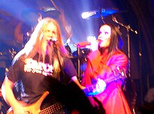 Image d'un homme blond jouant de la basse et d'une femme brune habillée de rouge chantant devant un public