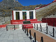 Mémorial en pierre rouge et blanc où sont fixées des plaques noires avec des listes de noms écrites en jaune, et en arrière-plan une crête rocheuse fortifiée.
