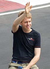 Head, arms and torso of a man in his early twenties with slightly tousled blond hair. He is waving with his right hand. He is wearing black T-shirt emblazoned with the Scuderia Toro Rosso on the left breast and beige trousers. Or shorts held up by a blue belt.