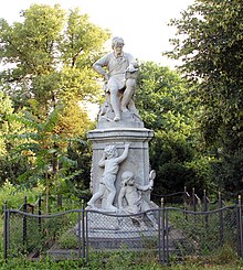 Photograph of a statue. A man, approximately life-size, is seated on a stool atop a pedestal. There are two children at the base of the pedestal. One child is standing, and appears to be tracing writing that is incised at the top of the pedestal. The other child is seated at the base, and is holding a hand mirror. The entire statue is about 12 feet (3.7 meters) high. It is situated in a park with trees in the background. An iron fence, about 1 meter high, encloses the base of the statue.