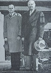 Two middle-aged men stand side-by-side wearing overcoats and holding their hats in their hands