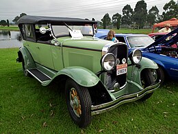 Limetkově zelené auto ve stylu 20. let 20. století s černým sklopným vrcholem směřujícím dopředu šikmo směrem k fotografii vpravo na trávě