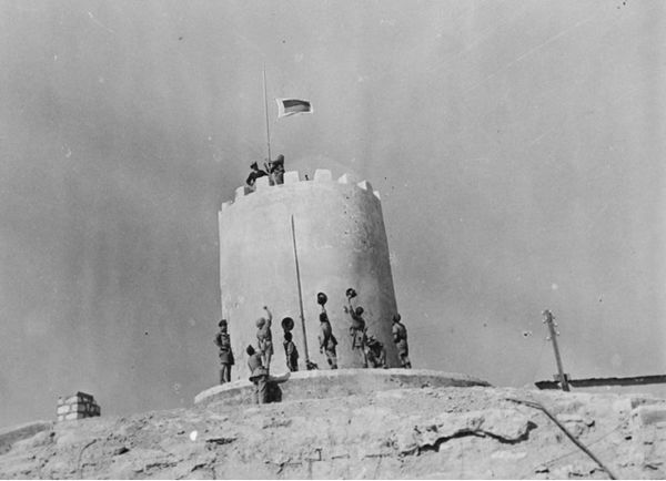 Soldiers from the 2/9th hoist a flag consisting of the battalion's unit colour patch over the recently captured Italian fort at Giarabub.