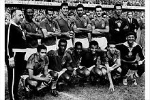 Schwarzweißes Gruppenfoto von dem ganzen Team. Sieben Männer stehen und sechs Männer hocken vor ihnen. Im Hintergrund ist ein volles Stadion zu sehen.