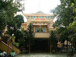 The front of the pagoda is cream colored and sits on a raised platform which is connected to the ground by a stairway. Chinese characters are above a set of ornate glass windows. A few people are sitting on the benches in the brick courtyard below, surrounded by many trees and shrubs. The roof is tiled brown and there is an unused flagpole at the front of the raise platform.