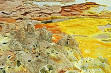 Formazioni saline e sulfuree sul Dallol. Si notano le concrezioni a forma di fungo (in secondo piano a destra), vasche di soda, colorate in giallo e arancio dallo zolfo (in primo piano a destra) e concrezioni spugnose (primo piano a sinistra).